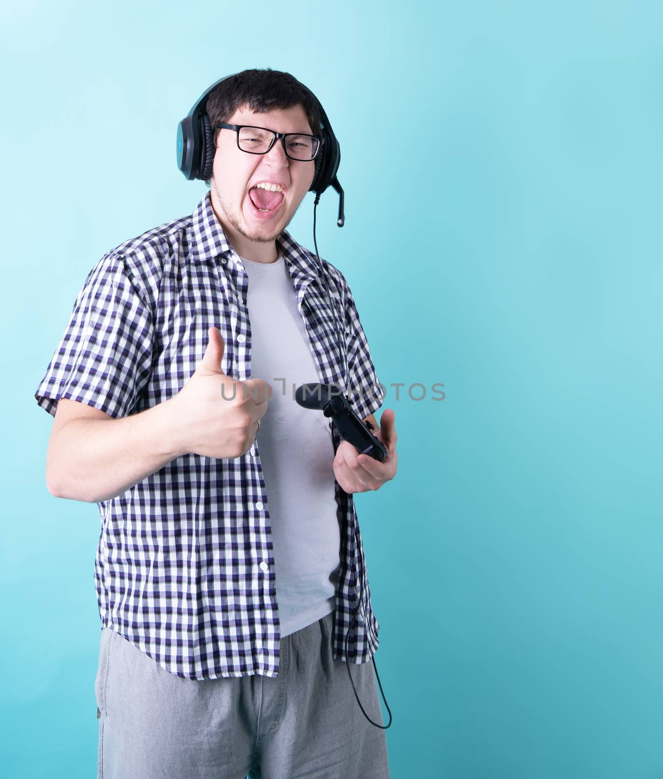 Funny young man playing video games holding a joystick showing thumb up isolated on blue background by Desperada