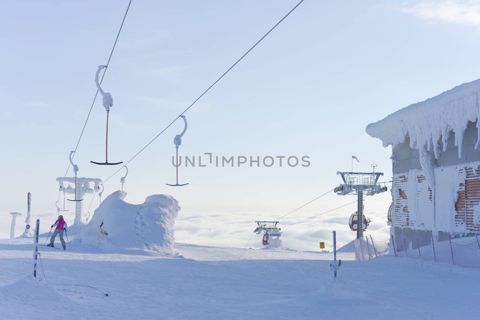 a single skier heads down a run having disembarked from ski lifts that rise up above the clouds into the pale blue sky. Very cold and snowy conditions.
