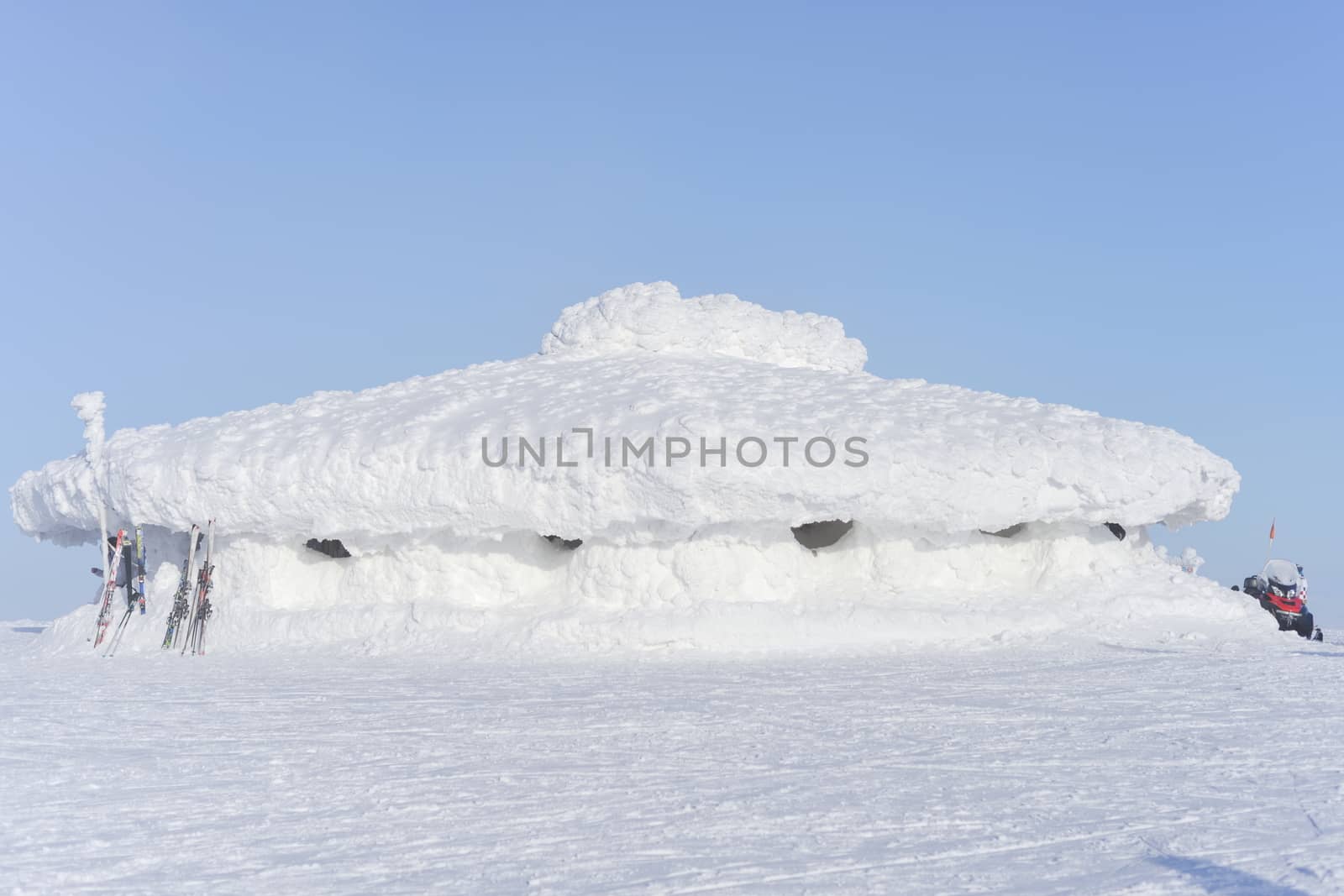 In the sunlight, this large, low lying building is covered in a very thick layer of bright white snow. Skis are rested against one end of the building.