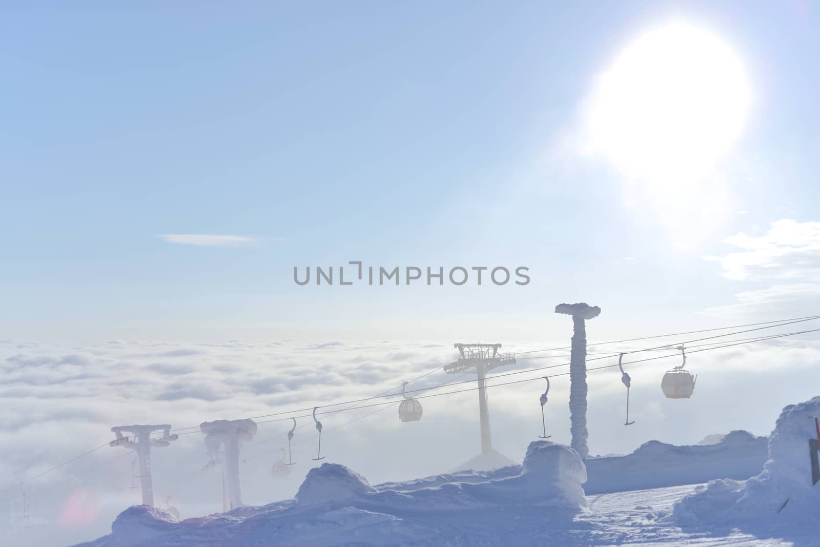 The sun flares from behind these gondola and T-bar ski lifts as they rise, frozen in snow and ice, from beneath the rolling clouds. Harsh and cold.