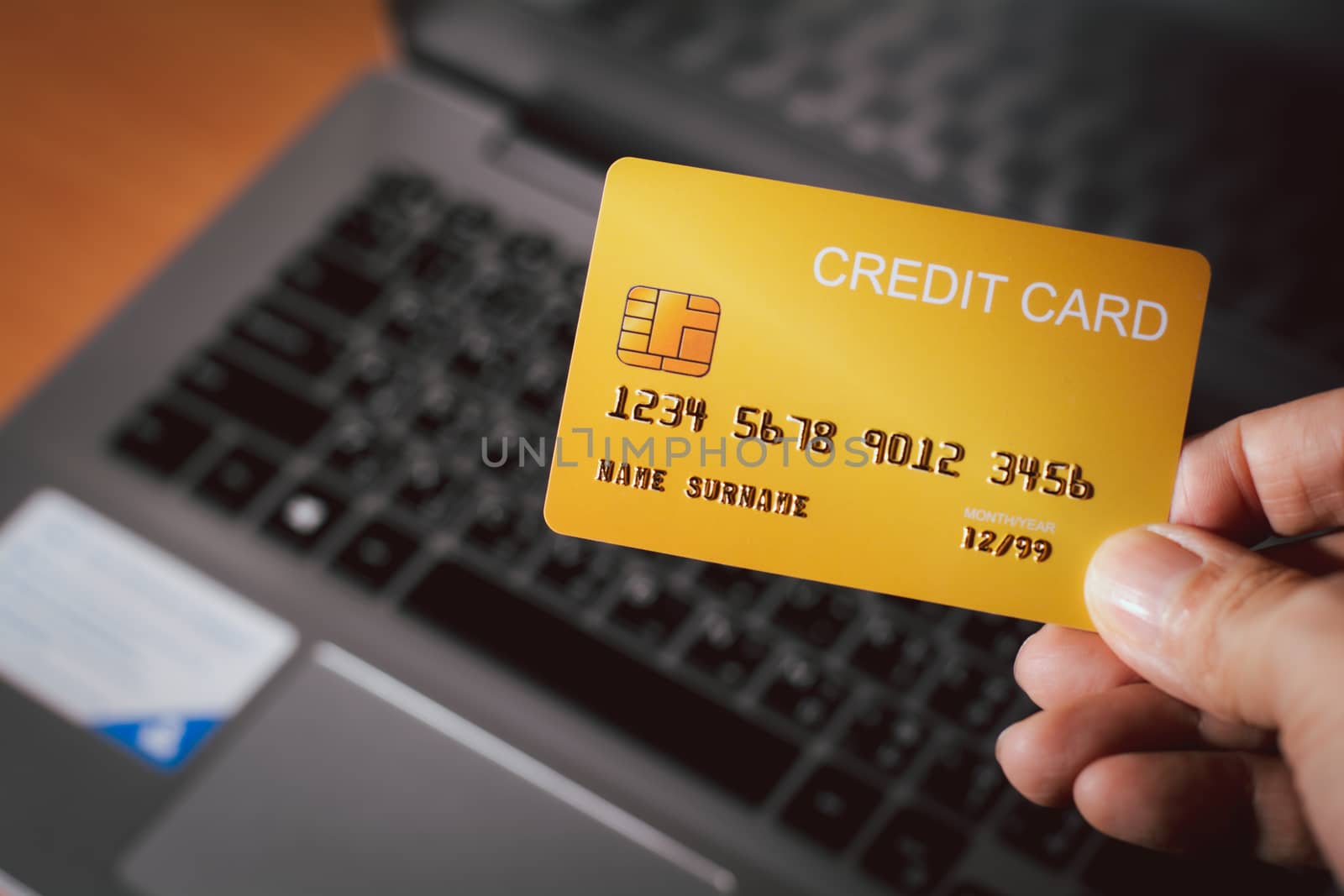 Shopping online and new normal concept. Hand holding yellow of credit card with blurry laptop background. Pay and buy online at home. 