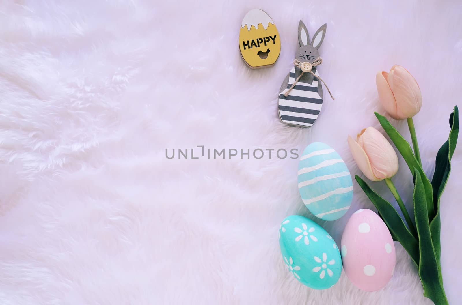 Happy Easter concept with wooden bunny and colorful easter eggs on white fur background and pink tulips. Top view with copy space 