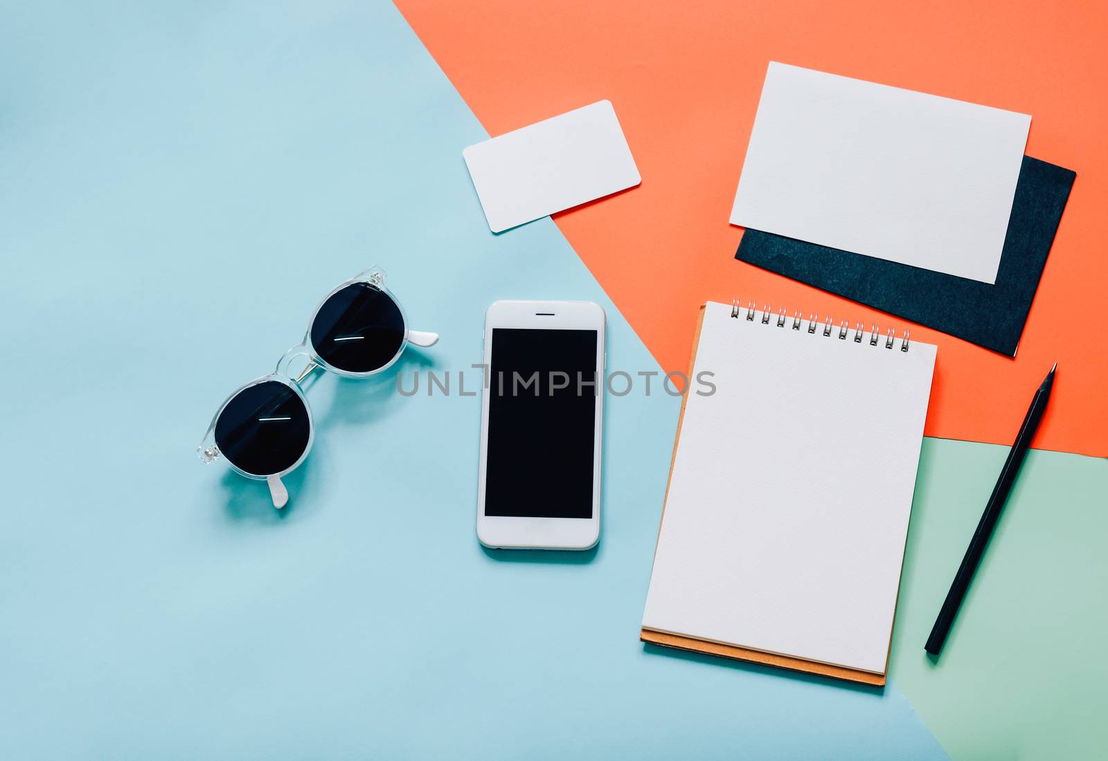 Creative flat lay style workspace desk with smartphone, blank envelope, sunglasses and blank notebook on modern colorful background