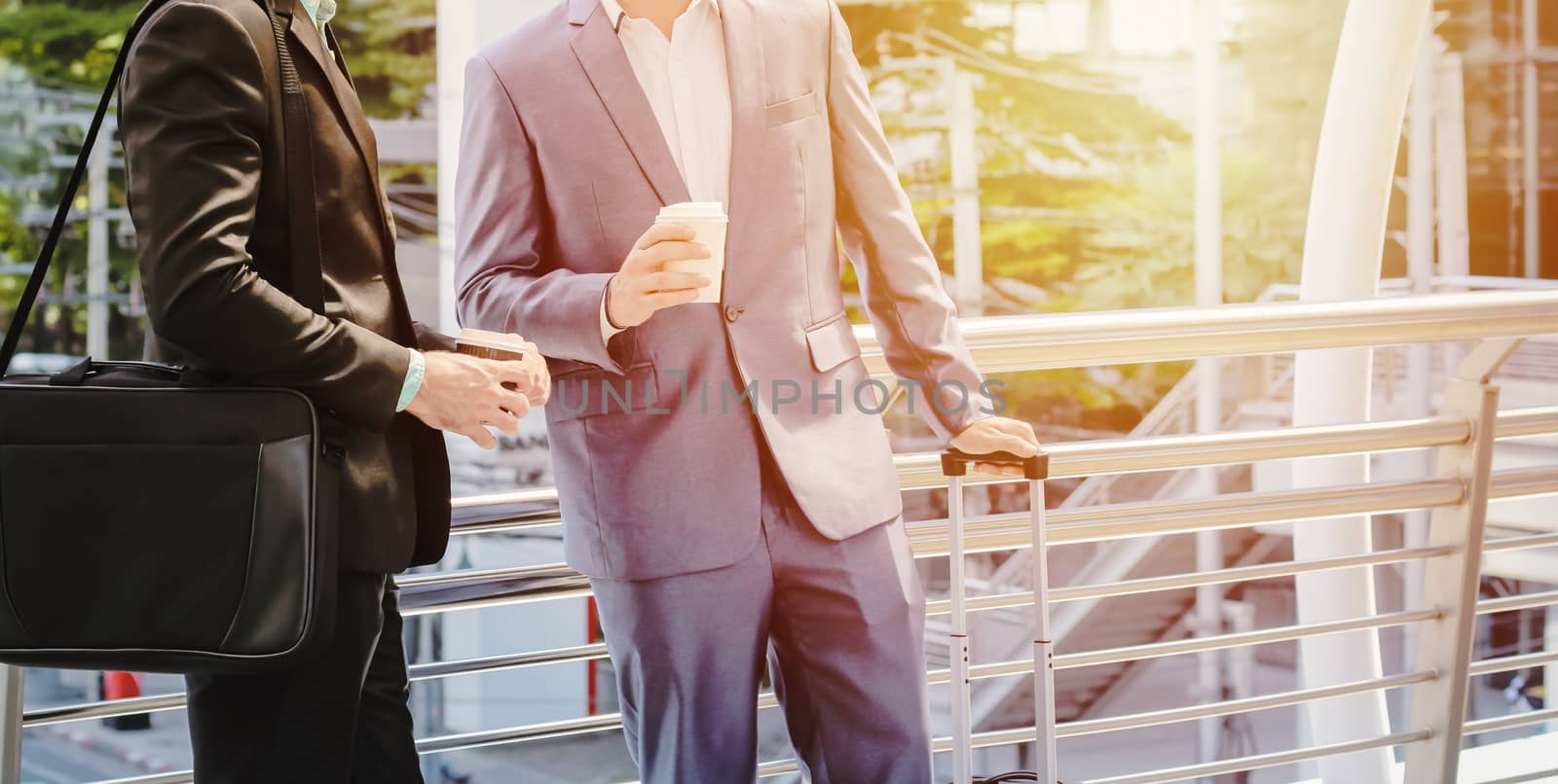 Businesspeople drinking a cup of coffee and holding luggage for business trip at sidewalk of the modern building with sunlight