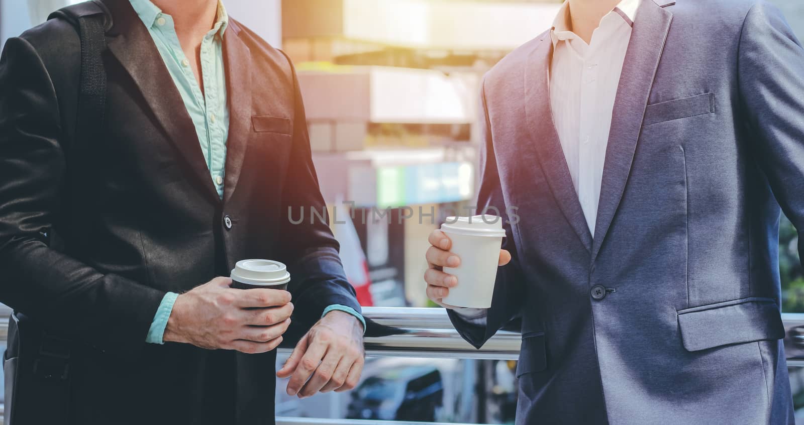 Midsection of two colleague businessmen using smartphone and holding coffee cup at the outside of modern office building with copy space and sunlight tone
