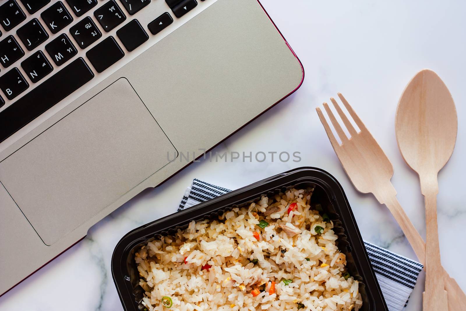 Delicious fried rice in lunch box on napery with wooden spoon and fork and computer laptop at workplace for ready to eat and food concept