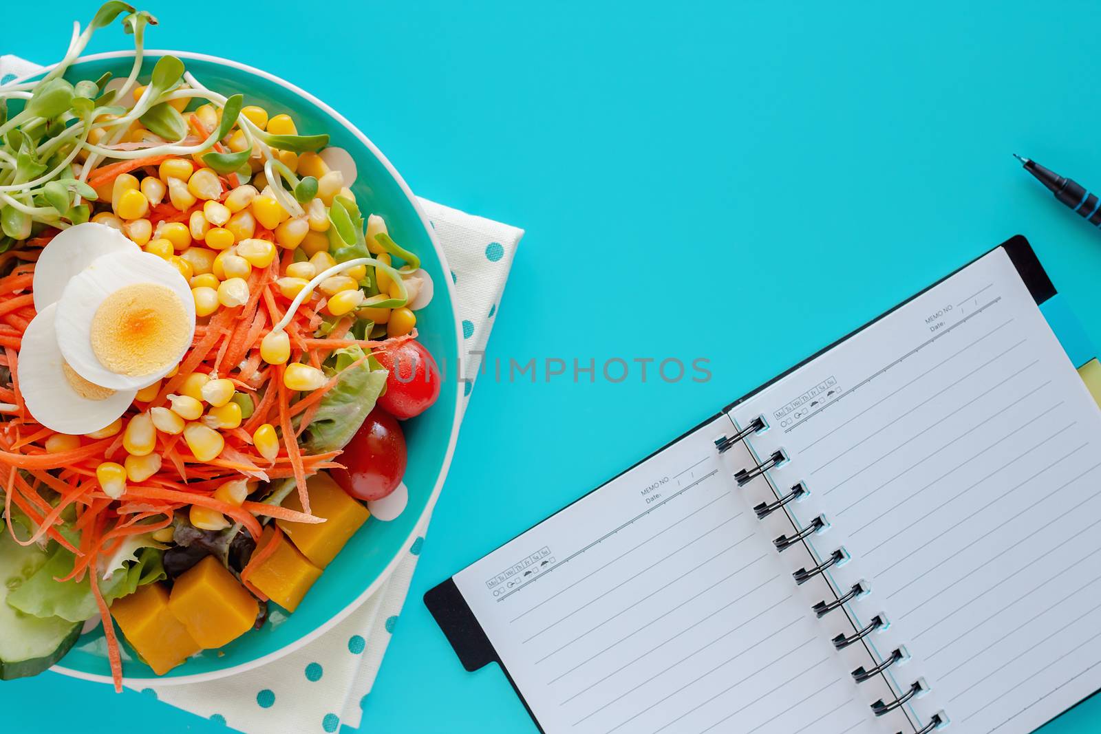 Fresh salad vegetable with boiled chicken egg, blank spiral notebook and a pen on blue background for healthy planning, eating and diet food concept