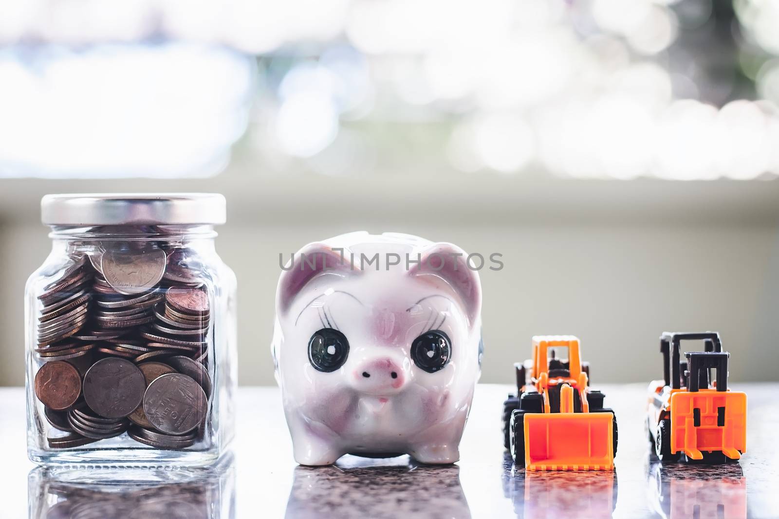 The glass bottle of coins and piggy bank with the yellow toy heavy machinery includes bulldozer and forklift against blurred background for saving money, investment, business and finance concep