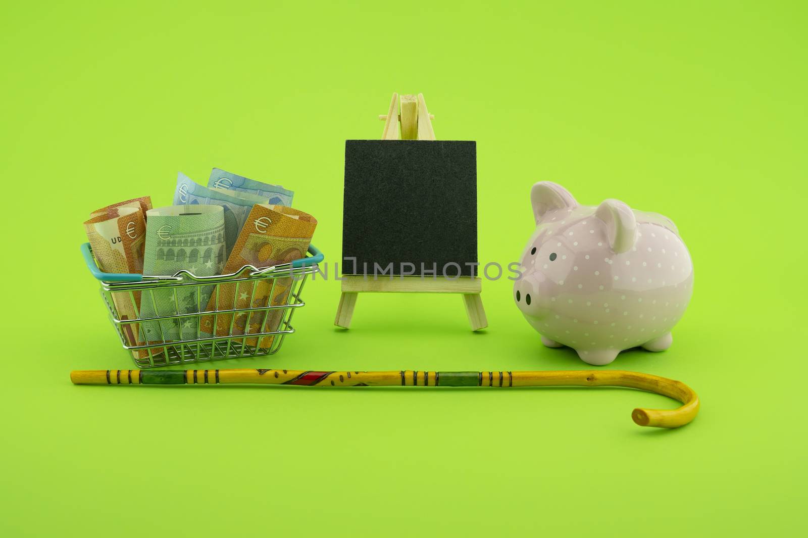Saving for old-age and retirement concept with shopping basket filled with cash banknotes, walking stick, chalkboard and piggy bank over a green background with copy space