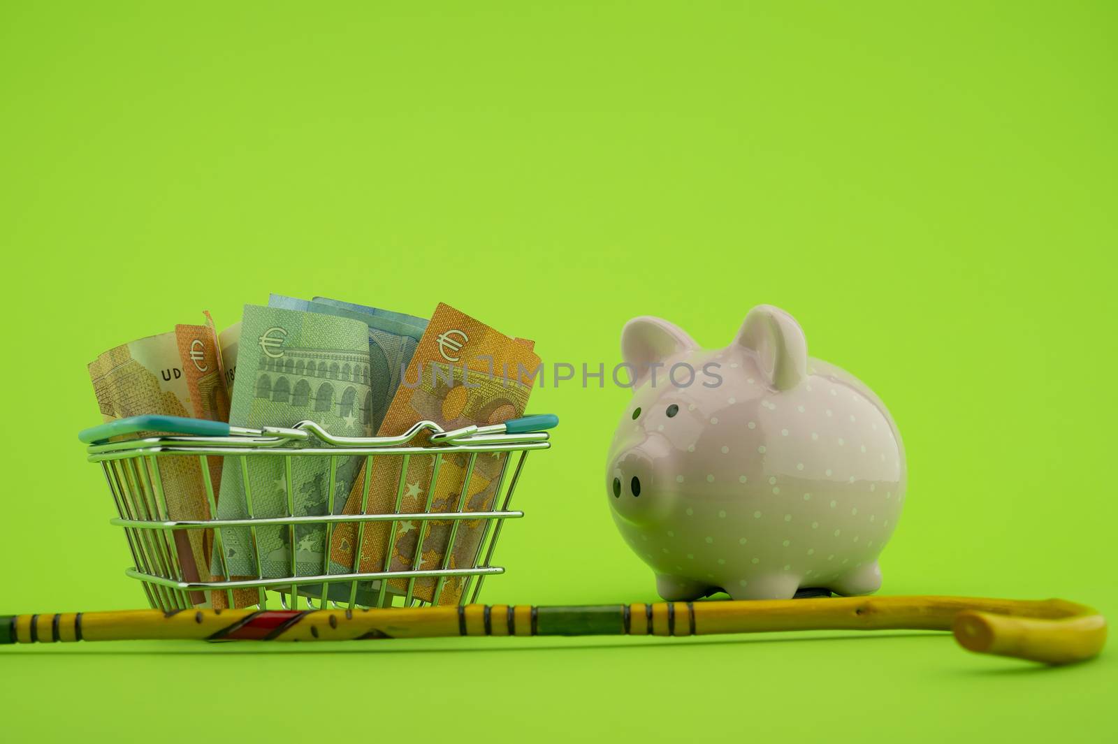 Saving for old-age and retirement concept with shopping basket filled with cash banknotes, walking stick and piggy bank over a green background with copy space