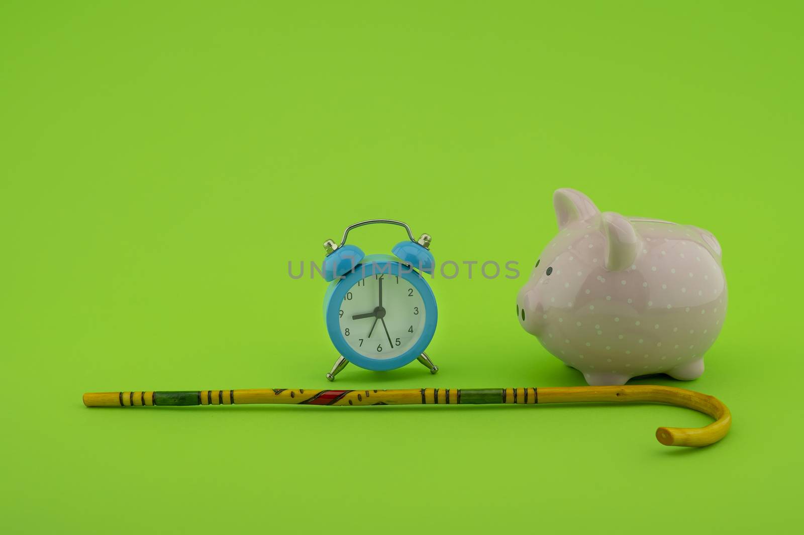 Saving for old-age and retirement concept with alarm clock, walking stick and piggy bank over a green background with copy space