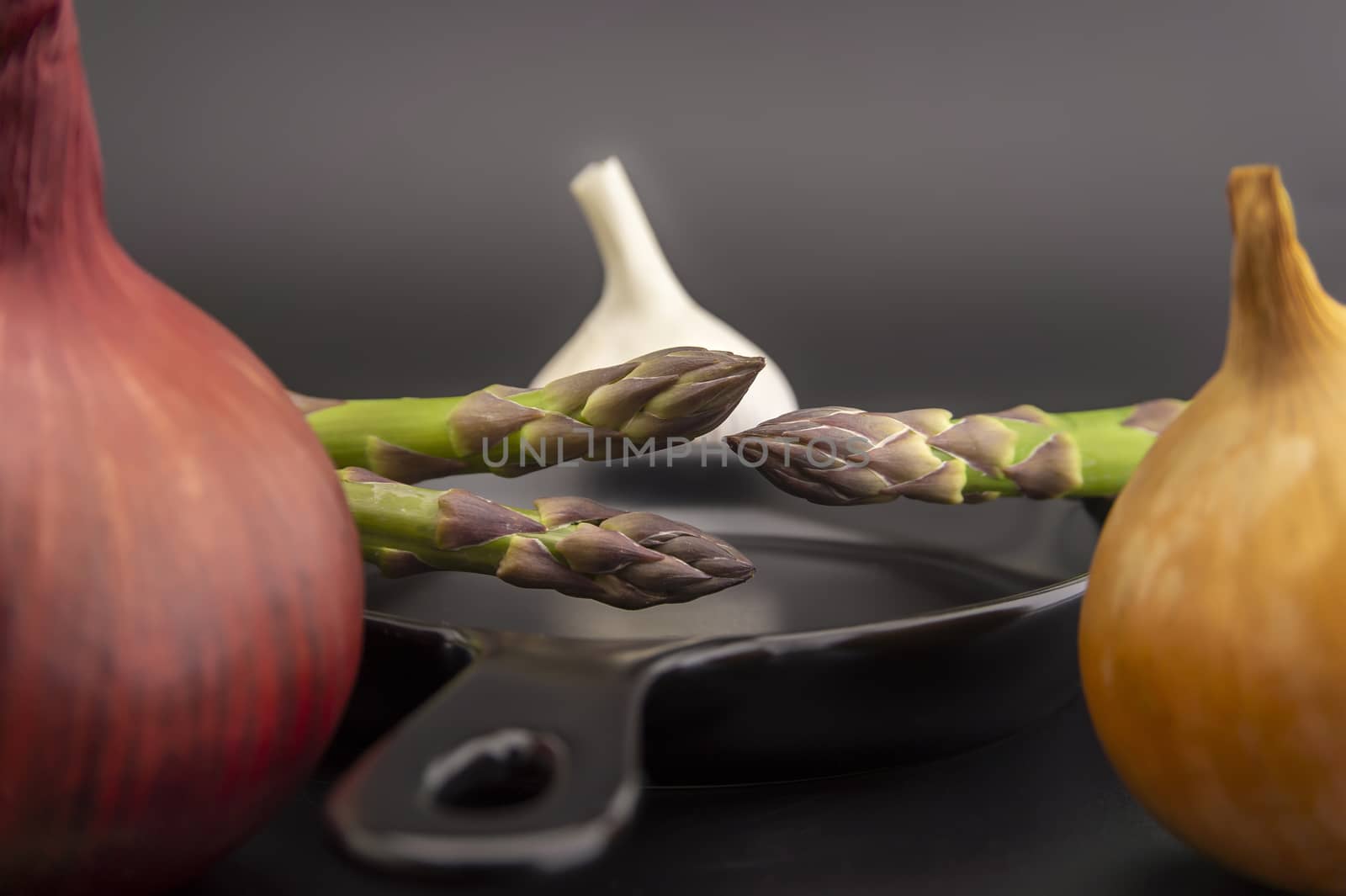 Fresh seasonal spring cooking ingredients surrounding a cast iron skillet with red and white onions, garlic bulb and green asparagus spears in a low angle view