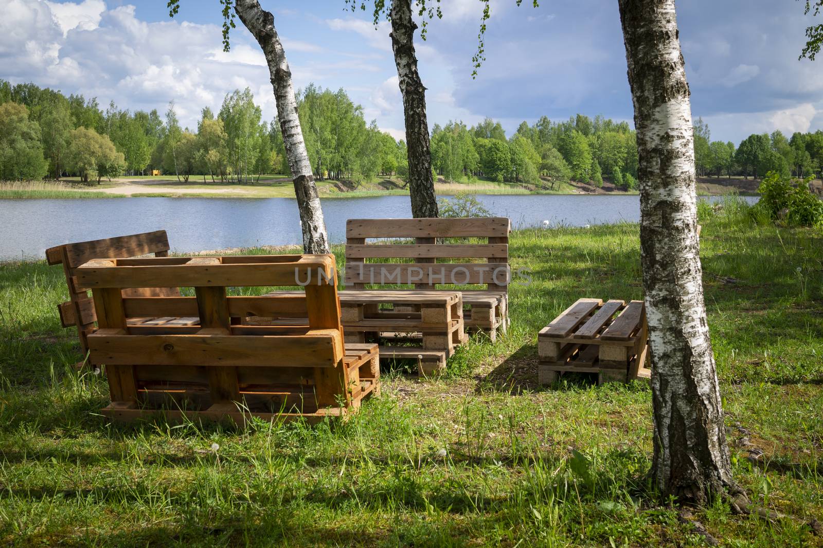 Rustic wooden table and benches made with pallets on the shore of a tranquil a lake surrounded by trees and greenery in summer sunshine