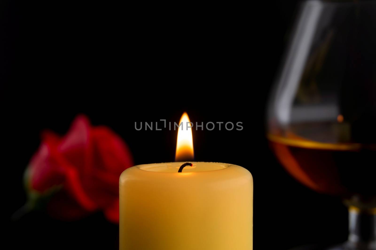 Single burning yellow candle in front of a glass of Cognac or Brandy and red roses against dark background