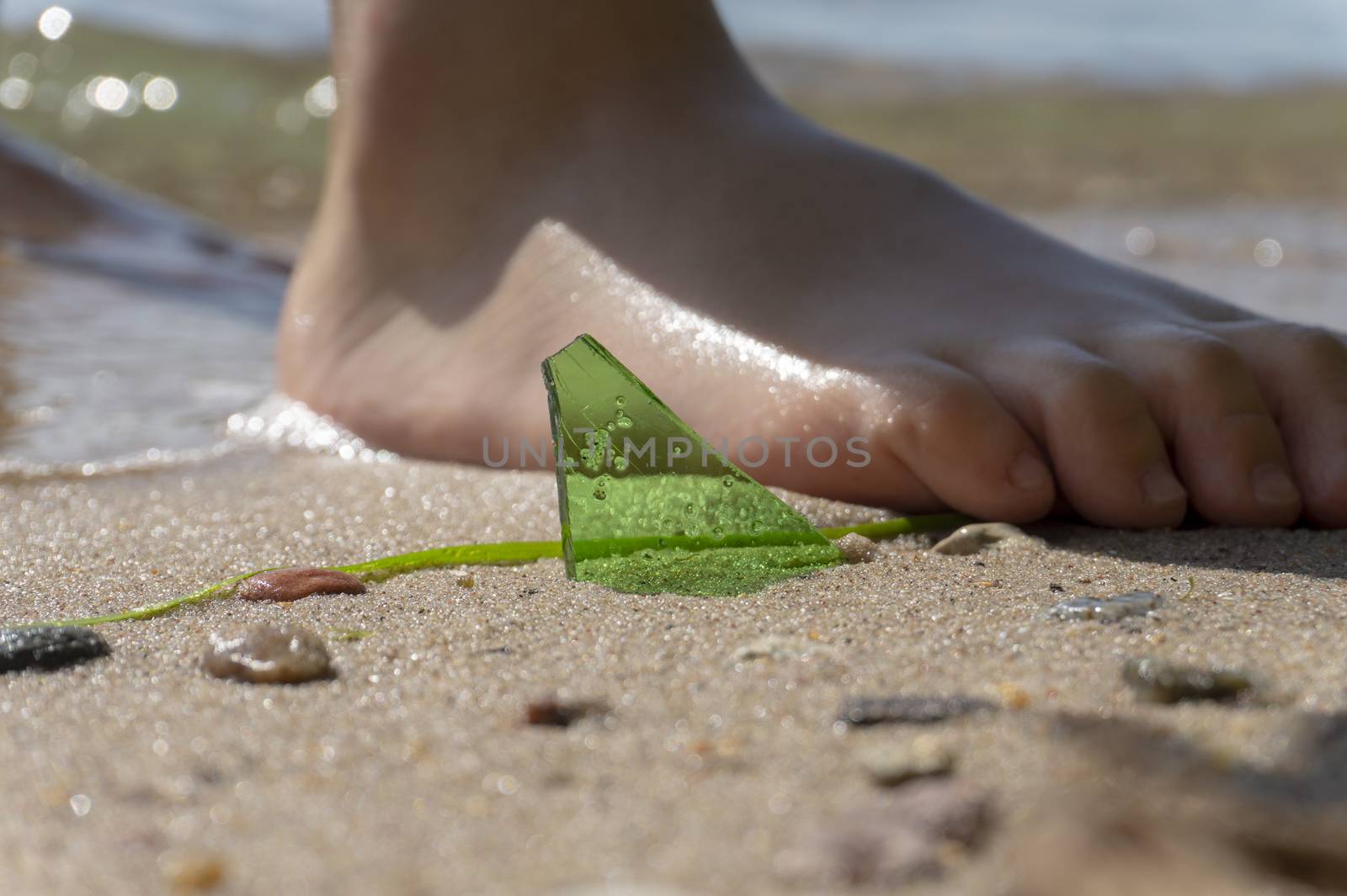 Danger of broken glass on a beach with a close up view of a shard of green glass protruding from the sand alongside the bare foot of a young person