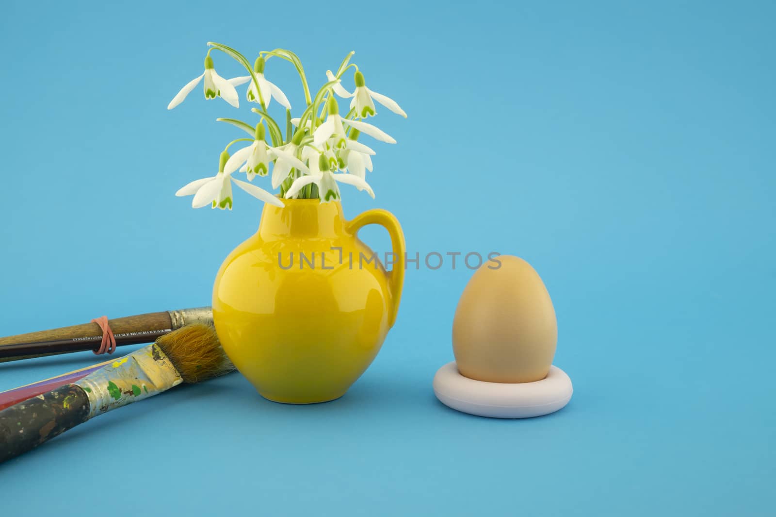 Easter egg decoration concept with paint brushes and yellow jug with fresh white spring flowers. Studio still life on blue background