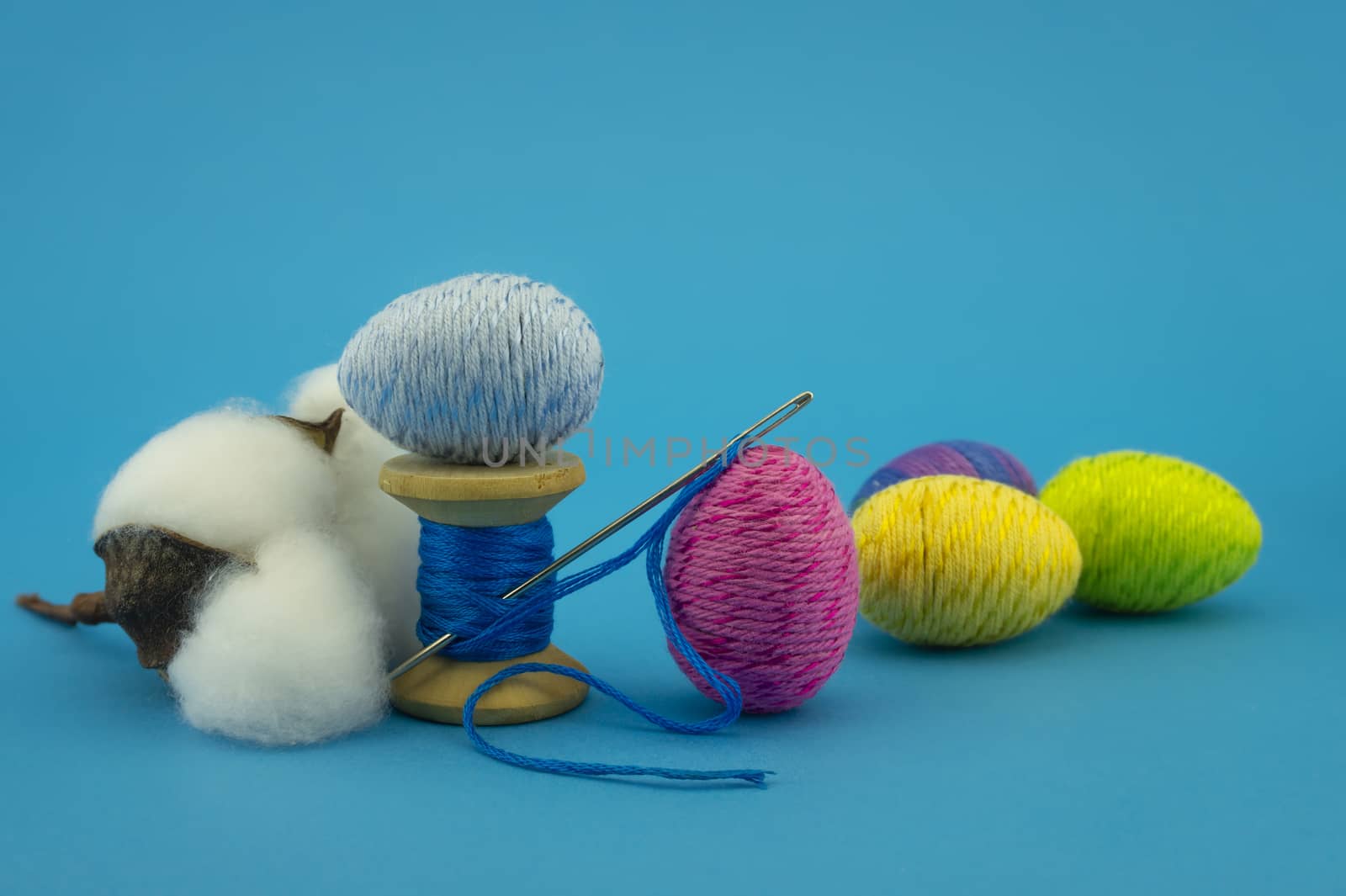 Handcrafted Easter decorations from natural cotton with a cotton boll off the plant alongside a reel of blue thread with needle and creative colorful eggs made from yarn over a blue background