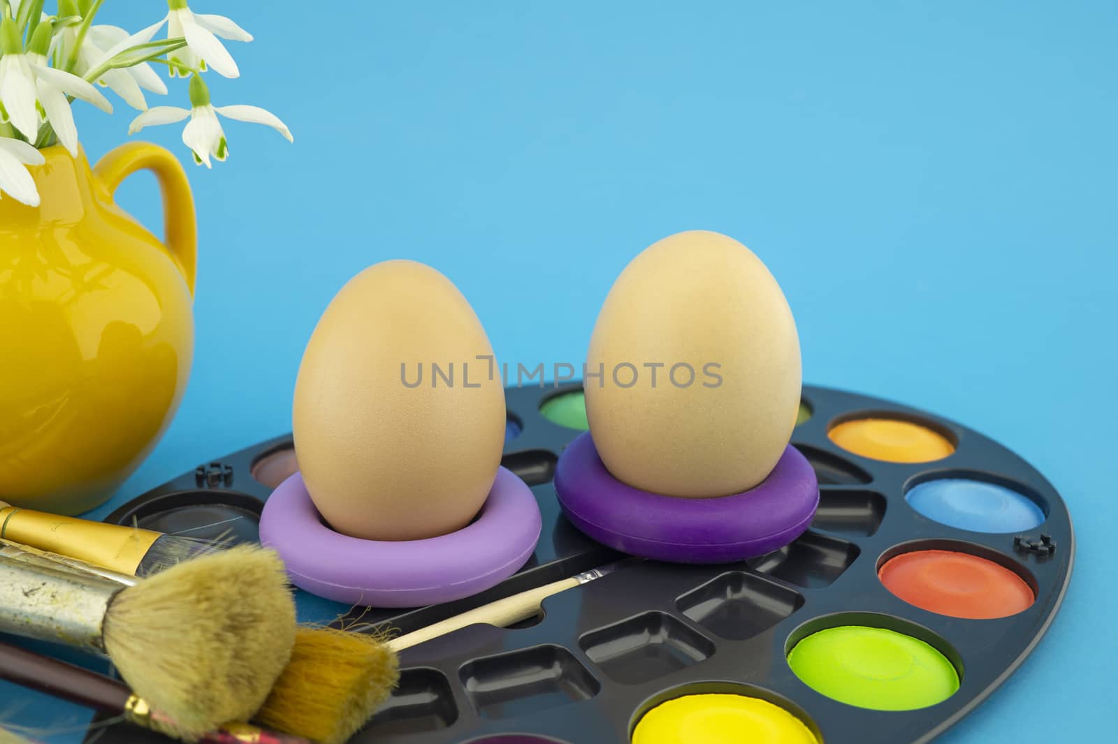 Easter egg decoration concept with paint brushes and set of watercolor paints in pallet, next to a yellow jug with fresh white spring flowers. Studio still life on blue background