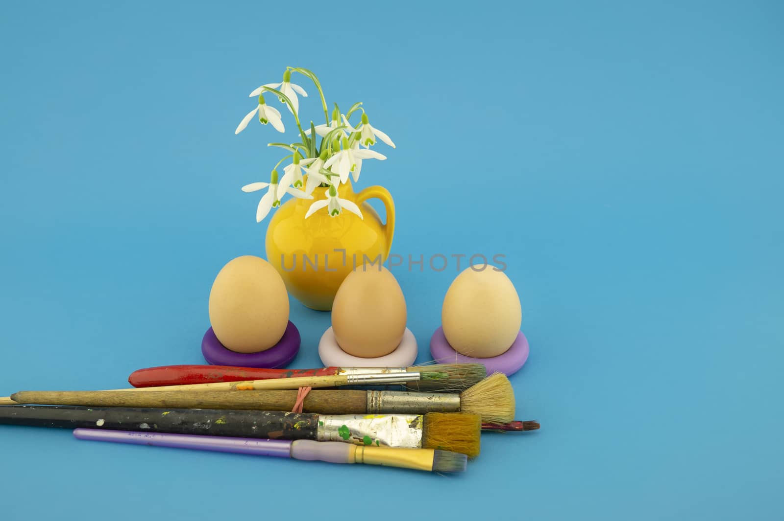 Easter egg decoration concept with paint brushes next to a yellow jug with fresh white spring flowers. Studio still life on blue background