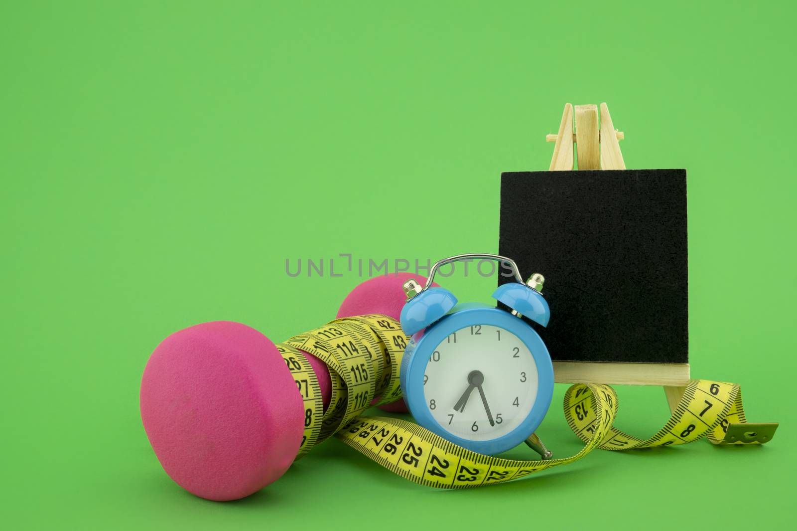 Starting healthy lifestyle concept. Still life with pink dumbbell, measuring tape, chalkboard and alarm clock on green background