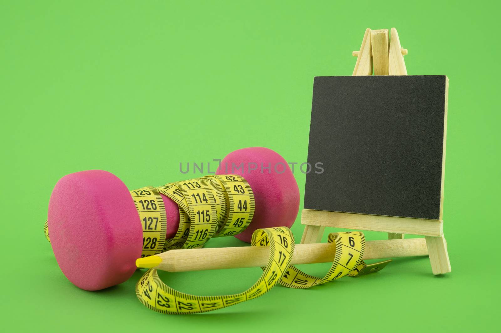 Starting healthy lifestyle concept. Still life with pink dumbbell, measuring tape, chalkboard and pencil on green background