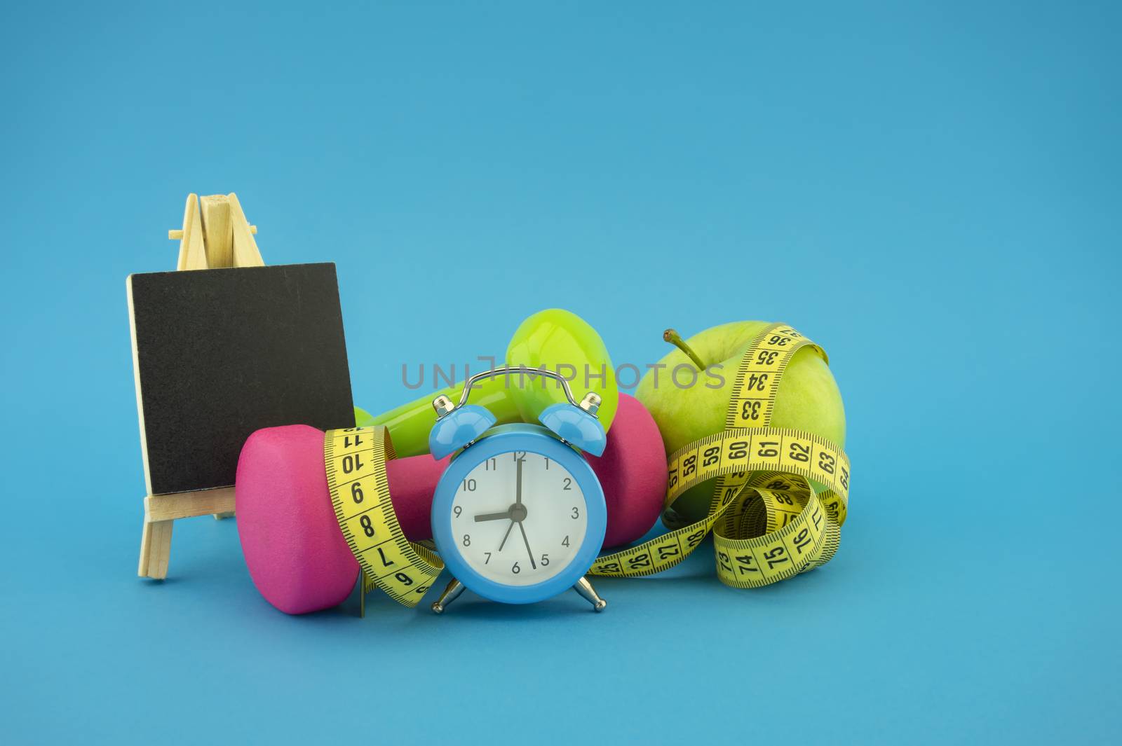 Starting healthy lifestyle concept. Still life with pink and green dumbbells, measuring tape, apple, chalkboard and alarm clock on green background