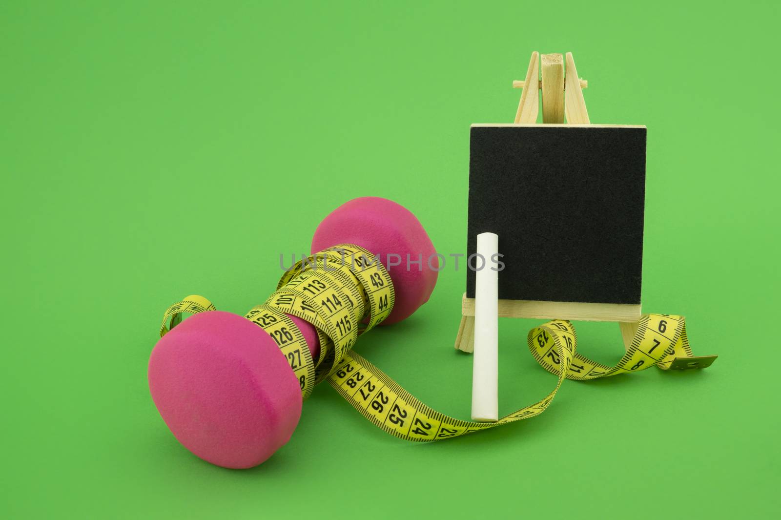 Starting healthy lifestyle concept. Still life with pink dumbbell, measuring tape, chalkboard and chalk on green background