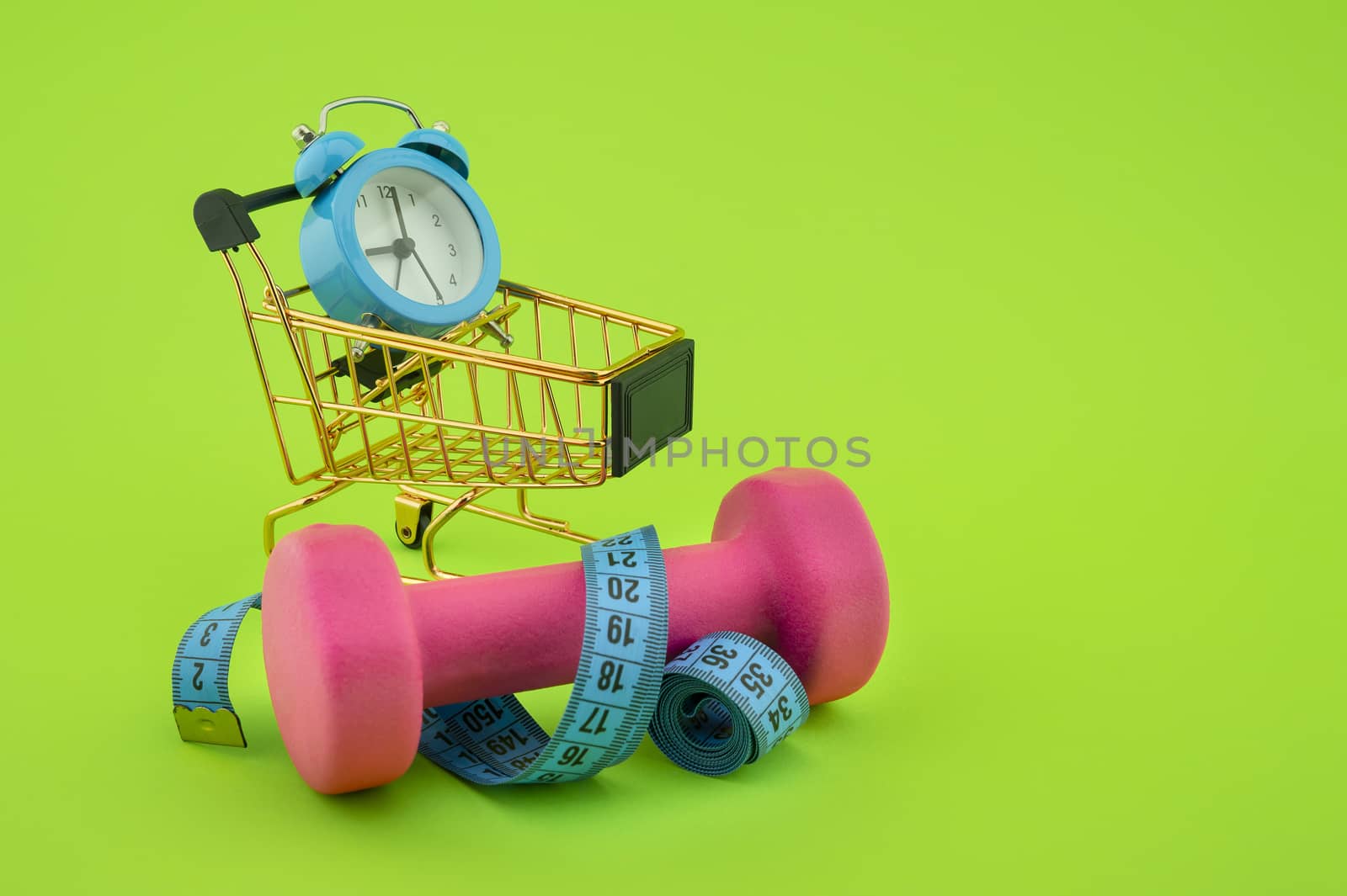 Going shopping for sports diet concept still life with dumbbells, measuring tape, alarm clock and wire shopping cart on green background