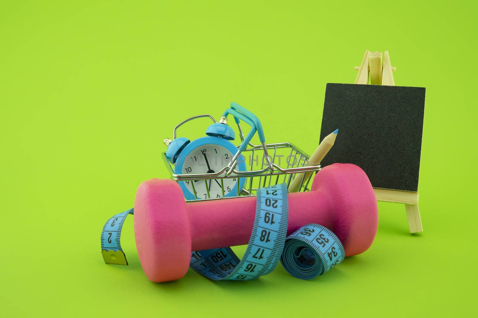 Starting healthy lifestyle concept. Still life with pink dumbbell, measuring tape, chalkboard and alarm clock on green background