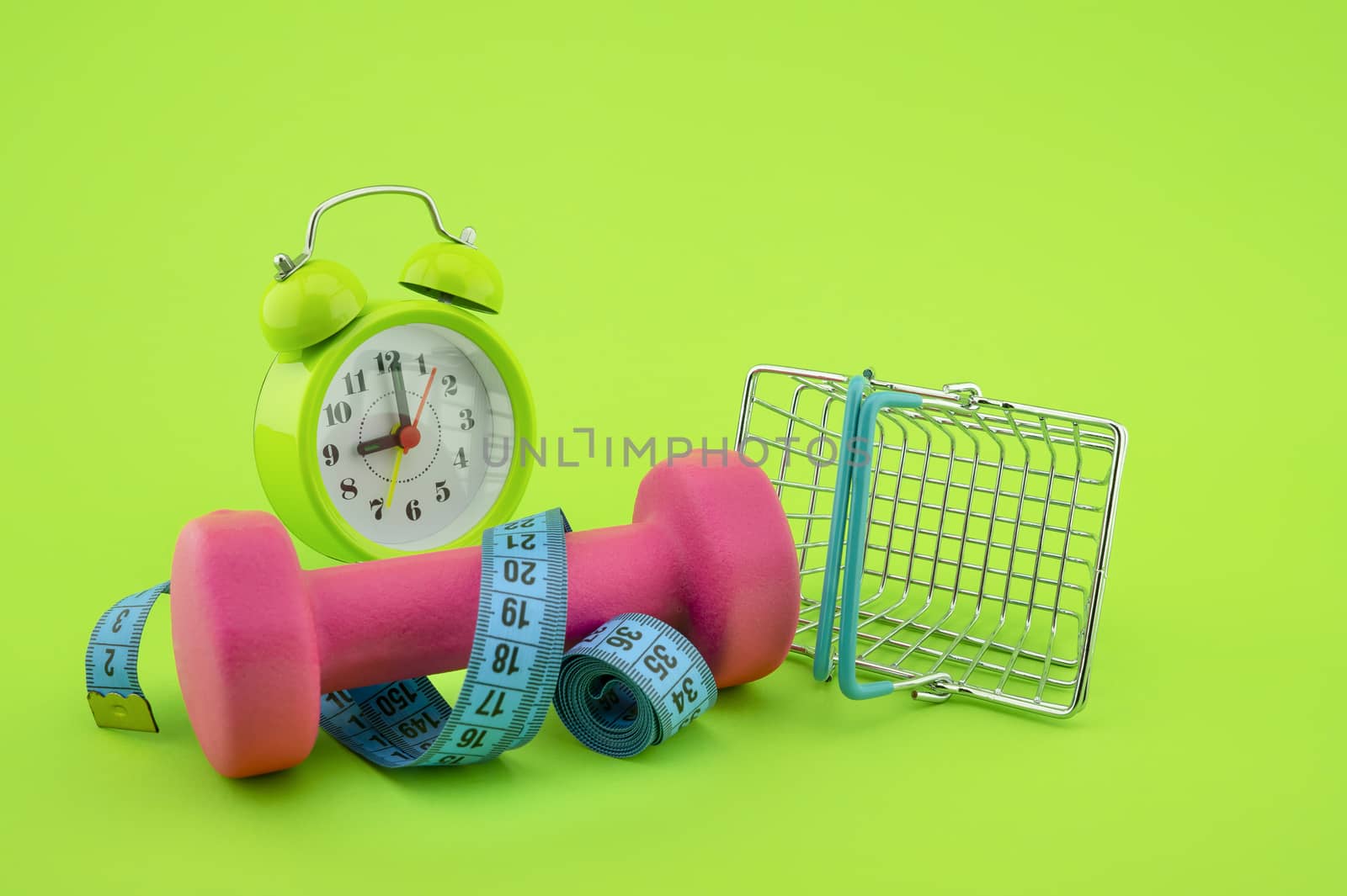 Going shopping for sports diet concept still life with dumbbells, measuring tape, alarm clock and wire shopping basket on green background