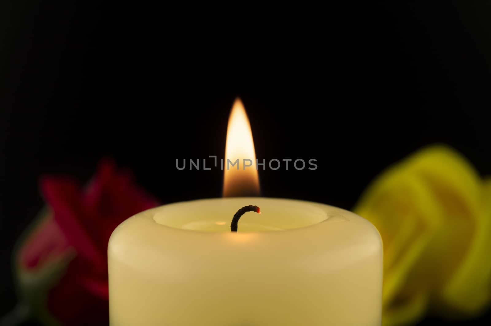 Single burning yellow candle next to red and yellow roses against dark background
