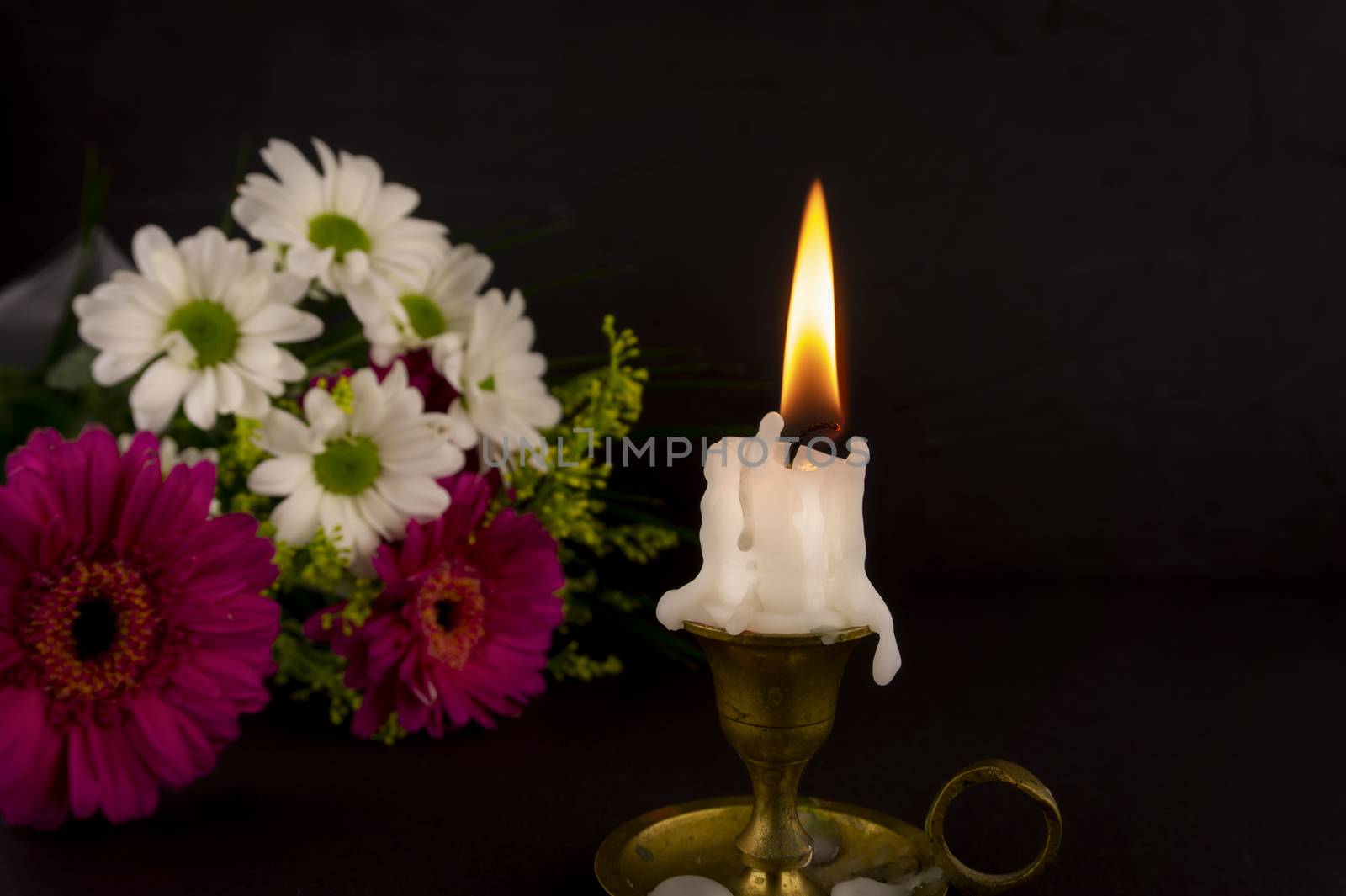 Short white candle burning on the floor in close-up among flowers against dark background.