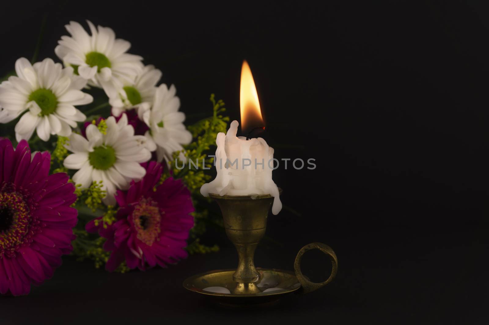 Short white candle burning on the floor in close-up among flowers against dark background.