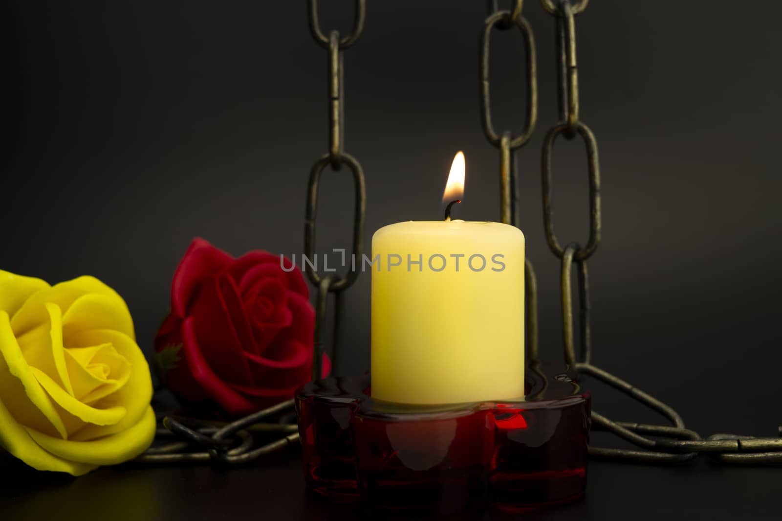 Dramatic still life with close-up of a burning yellow candle next to a metallic chain and two roses