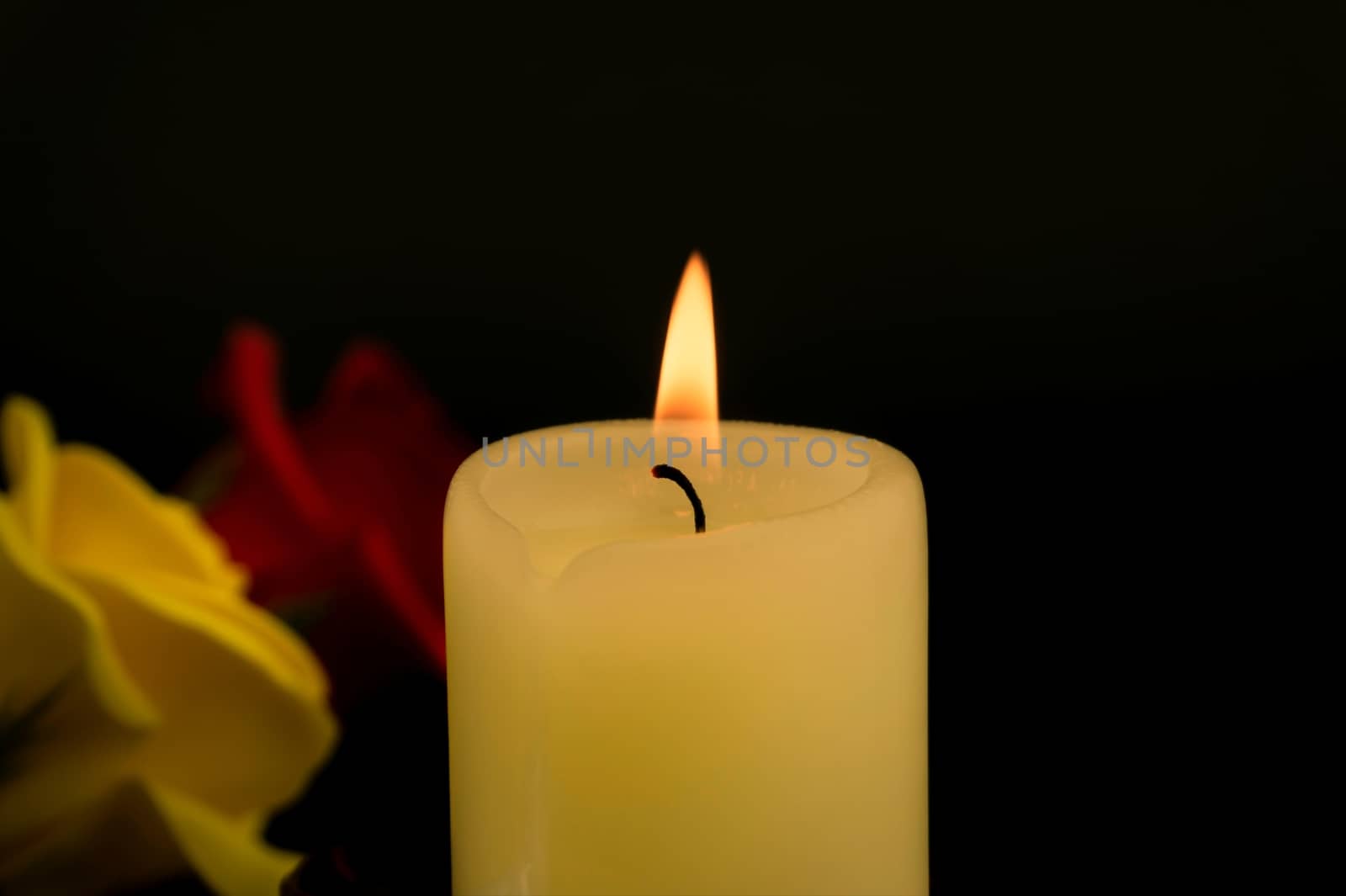 Single burning yellow candle next to red and yellow roses against dark background