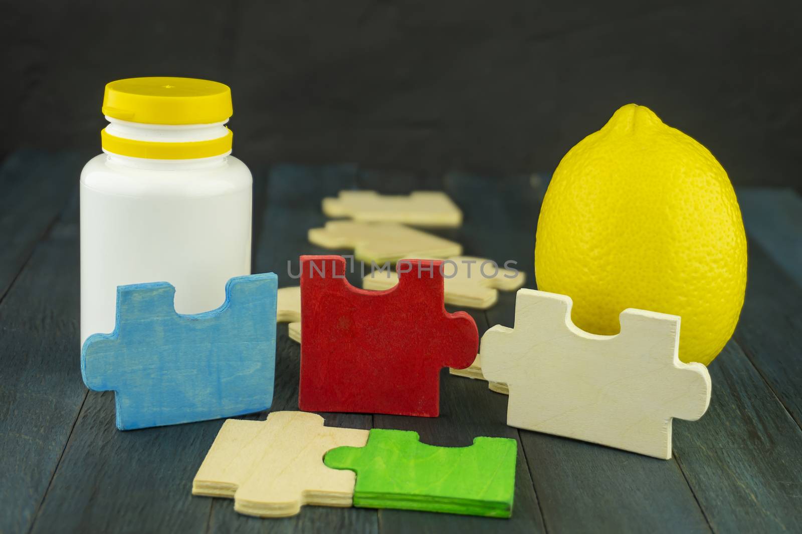 Plastic bottle of vitamins and whole lemon in a still life with colorful puzzles, viewed in close-up on dark wooden table. Healthy nutrition concept