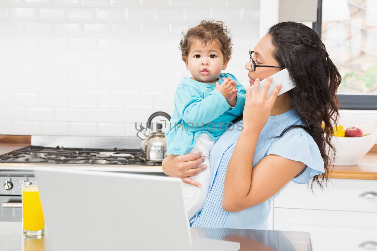 Smiling brunette holding her baby and using laptop on phone call in the kitchen