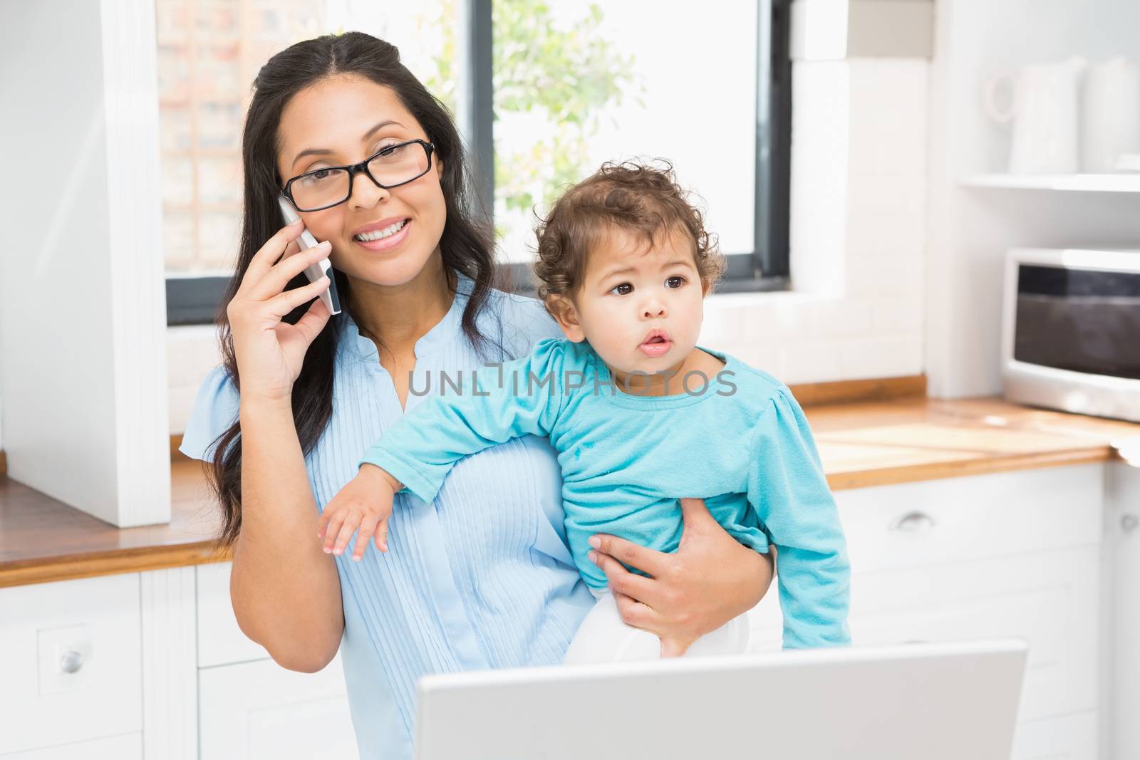 Smiling brunette holding her baby and using laptop on phone call in the kitchen