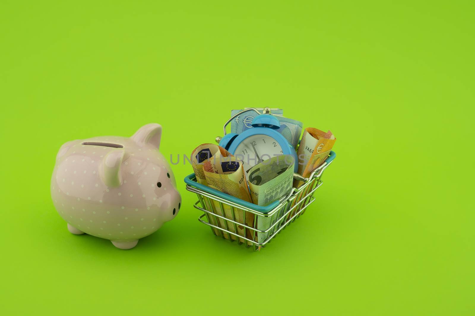 Shopping concept with ceramic piggy bank, basket filled with cash banknotes and alarm clock over a green background