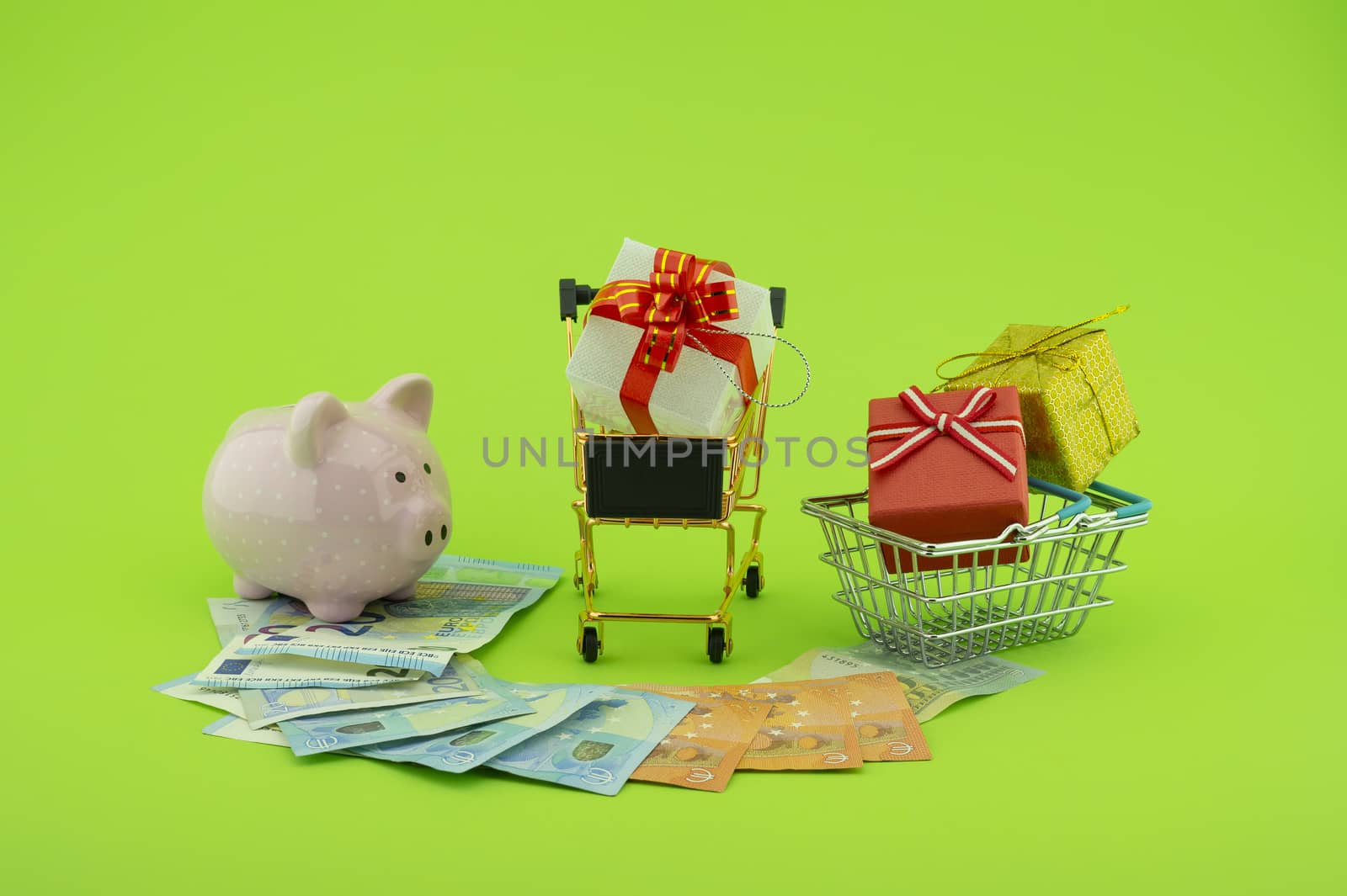 Cost of shopping or spending savings concept with banknotes scattered on a green background with a piggy bank, small wire shopping basket and cart with gifts with decorative red ribbons