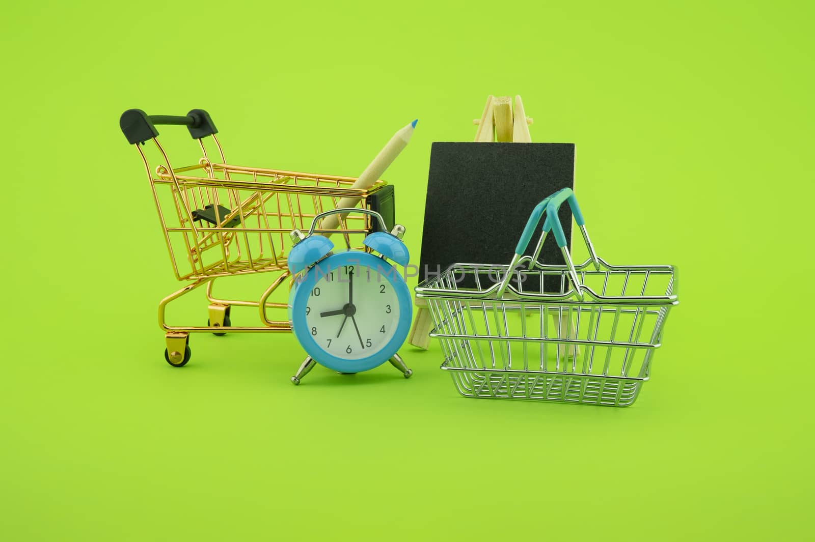Shopping concept with shopping cart, basket and alarm clock, small chalk board over a green background