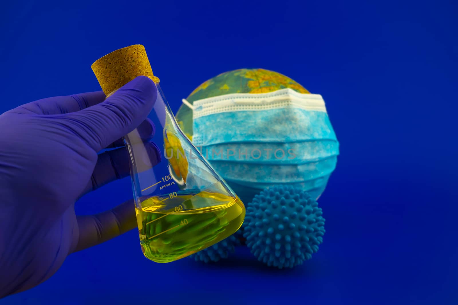 Coronavirus or Covid-19 still life with a gloved hand holding a laboratory flask with liquid or a possible vaccine in front of a globe with face mask and two virus molecules in blue toned theme