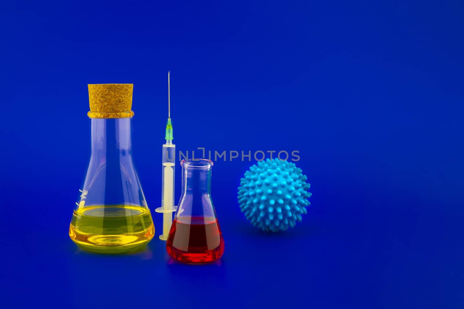 Concept of research for a Covid-19 vaccine with a conical lab flasks with yellow and red solutions alongside a hypodermic syringe and blue virus molecules over a blue background with copy space