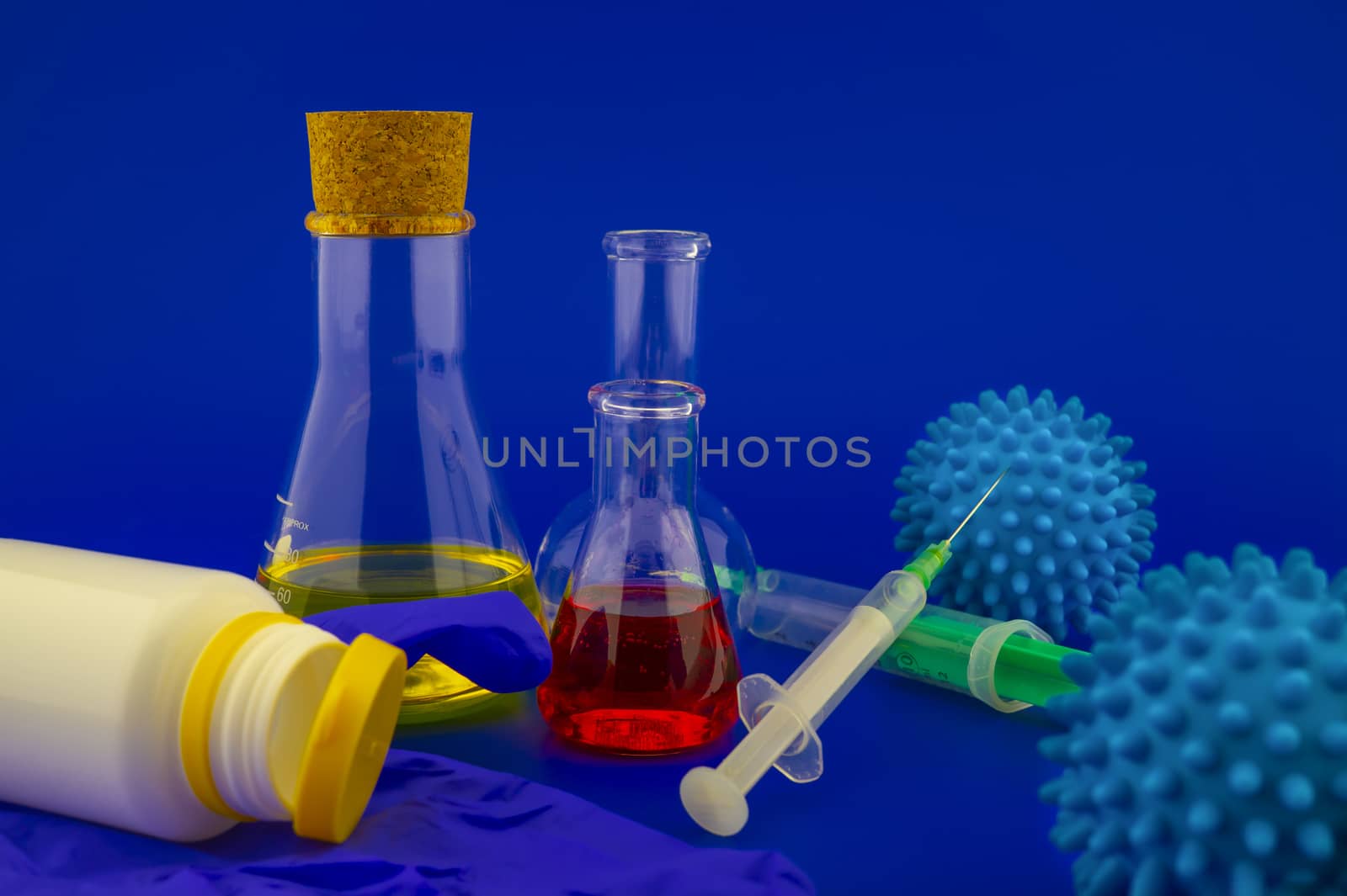 Concept of research for a virus vaccine with a conical lab flasks with yellow and red solutions alongside a hypodermic syringe and blue virus molecules over a blue background with copy space