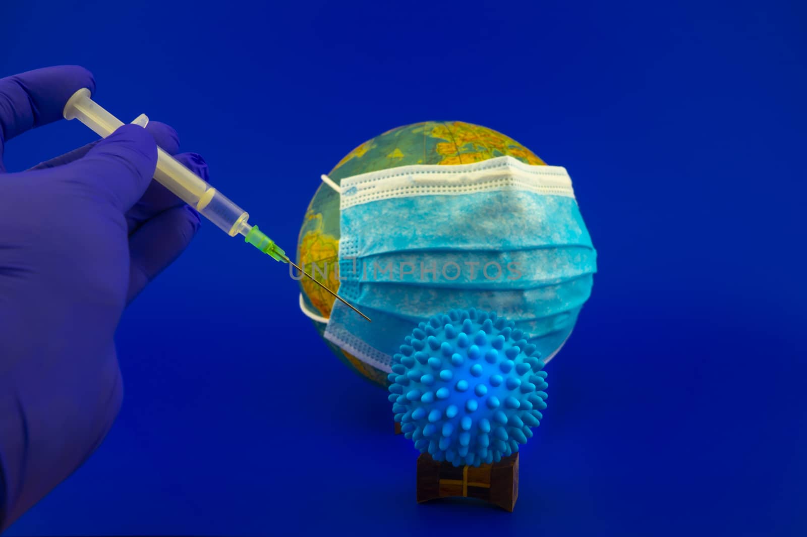 Coronavirus or Covid-19 pandemic concept with gloved hand holding a hypodermic syringe in front of a globe with protective face mask and two virus molecules over blue