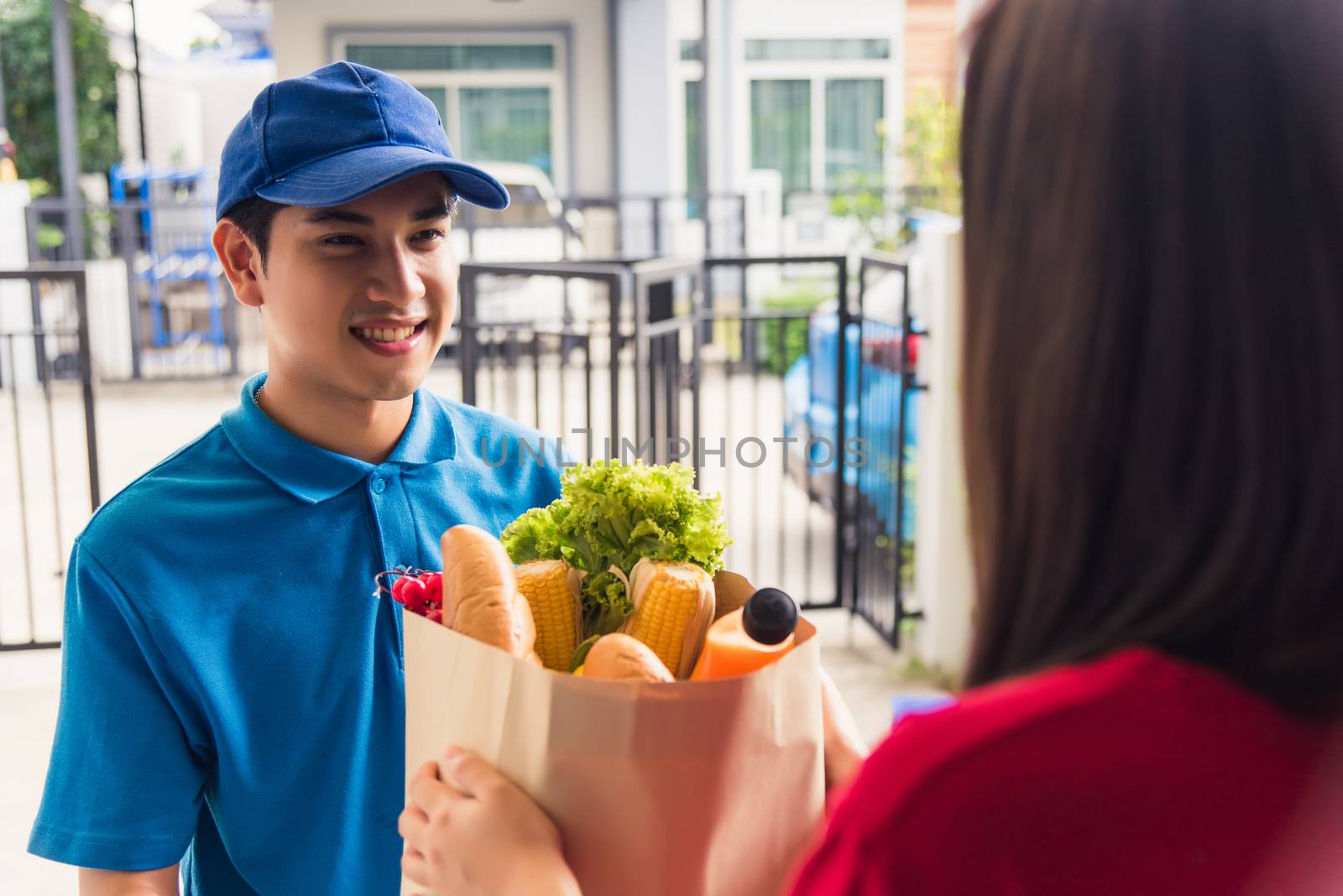 Asian young delivery man in uniform making grocery fast service giving fresh food in paper bag to woman customer receiving at house door under pandemic coronavirus, Back to new normal concept