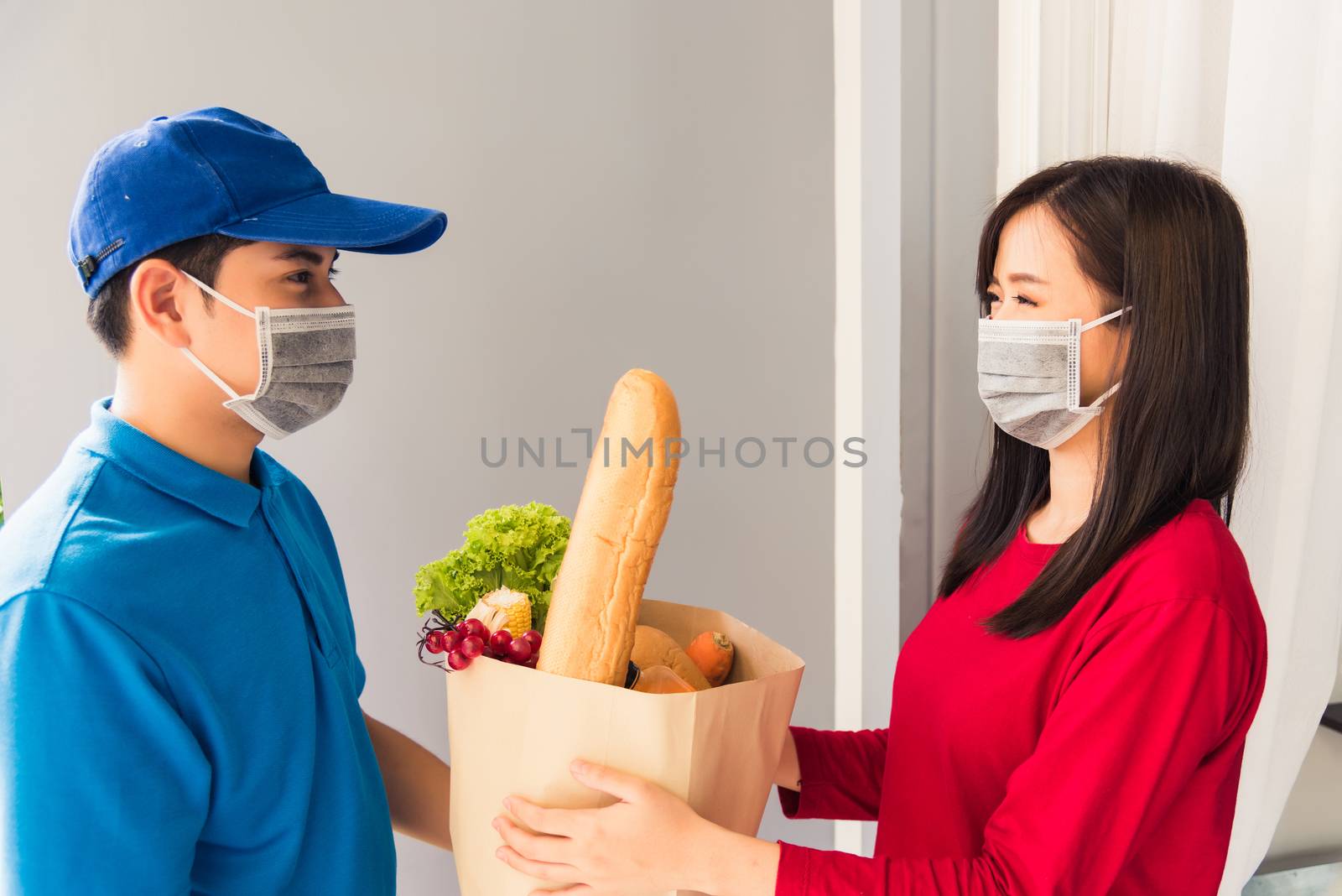 Asian young delivery man in uniform wear protective face mask he making grocery service giving fresh food to woman customer receiving front house under pandemic coronavirus, Back to new normal concept