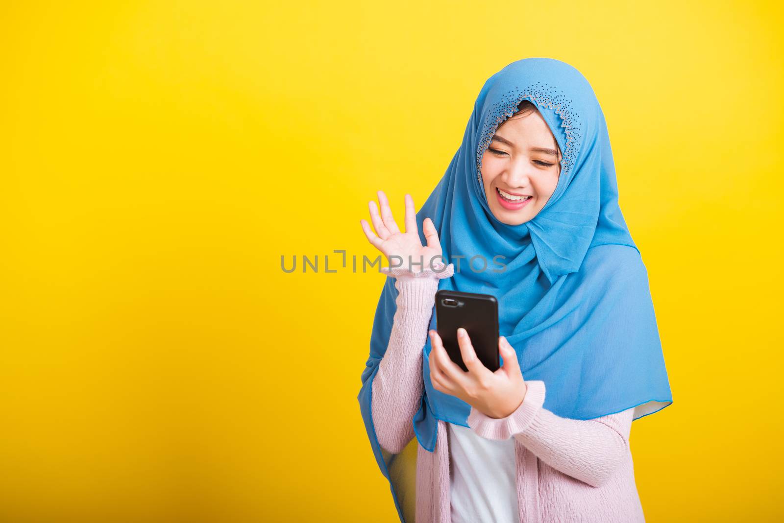 Asian Muslim Arab, Portrait of happy beautiful young woman Islam religious wear veil hijab funny smile she selfie or video call mobile smart phone raise hand say hello isolated on yellow background