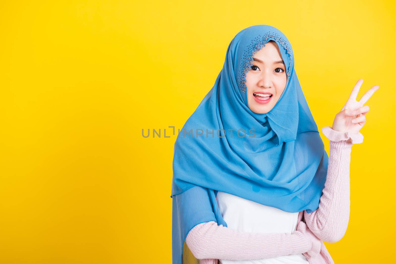 Asian Muslim Arab, Portrait of happy beautiful young woman Islam religious wear veil hijab smile she show gesture hand two finger V-sign symbol near eyes isolated yellow background
