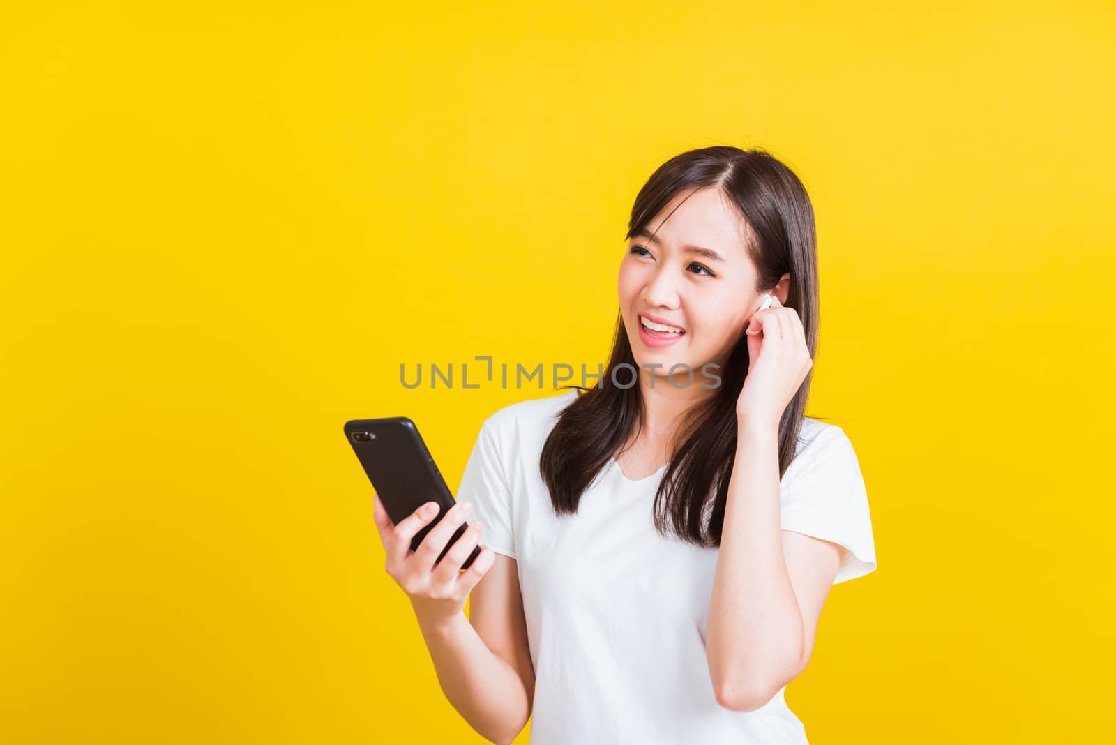 Portrait Asian of a happy beautiful young woman holding a mobile smart phone and wearing wireless headphones listening to music from smartphone studio shot isolated on yellow background